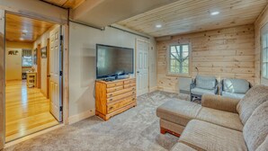 A log cabin interior with a wooden ceiling, a television on a dresser, a sofa, and two chairs. - Private River Access & Mtn Views (Woodstock)