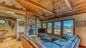 A cozy living room with a stone fireplace, leather sofa, and large windows offering a view of the outdoors. - Private River Access & Mtn Views (Woodstock)