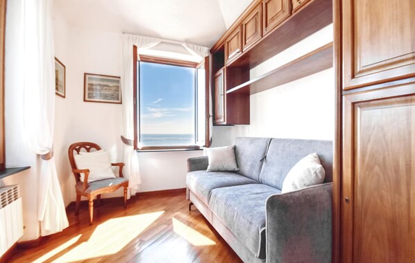 A cozy living room with a wooden floor, a grey sofa, a wooden chair, and a window offering a view of the sea.