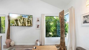 A modern living room with a large flat-screen TV, a wooden coffee table, and a sofa. There are two windows with curtains, and a large tree trunk decoration is visible. - Complex | Theater, Pickleball & Pool in DT Phoenix (Phoenix)