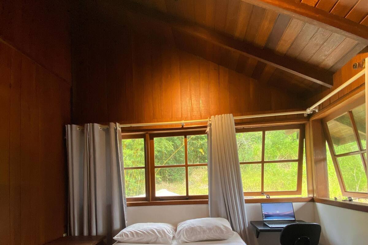 Une chambre avec un lit, un bureau avec un ordinateur portable, et une vue sur la verdure par la fenêtre.
