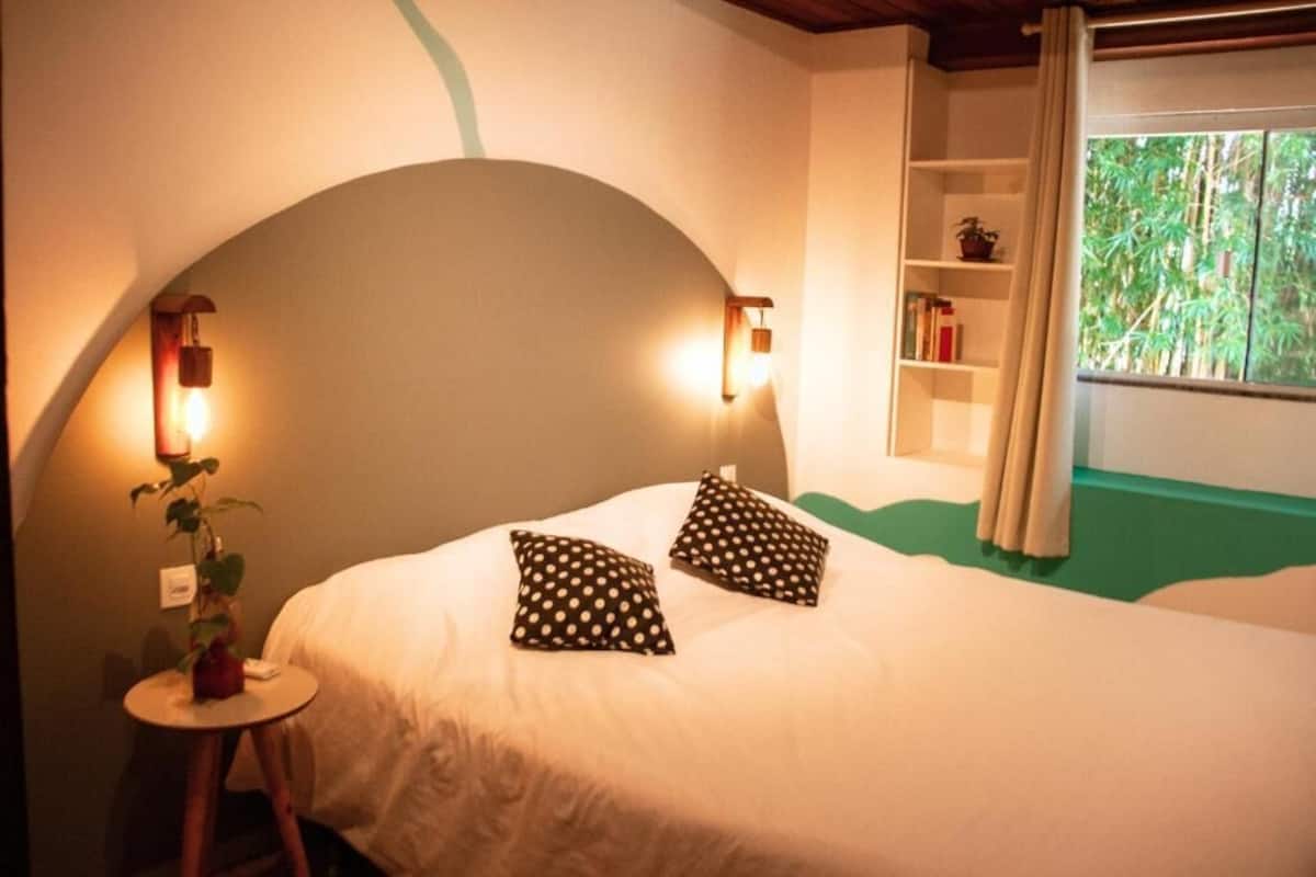 Une chambre à coucher avec un plafond incurvé, deux lampes de chevet, un lit avec une literie blanche, deux coussins à pois, une petite table de chevet avec une plante, et une fenêtre donnant sur un paysage verdoyant.