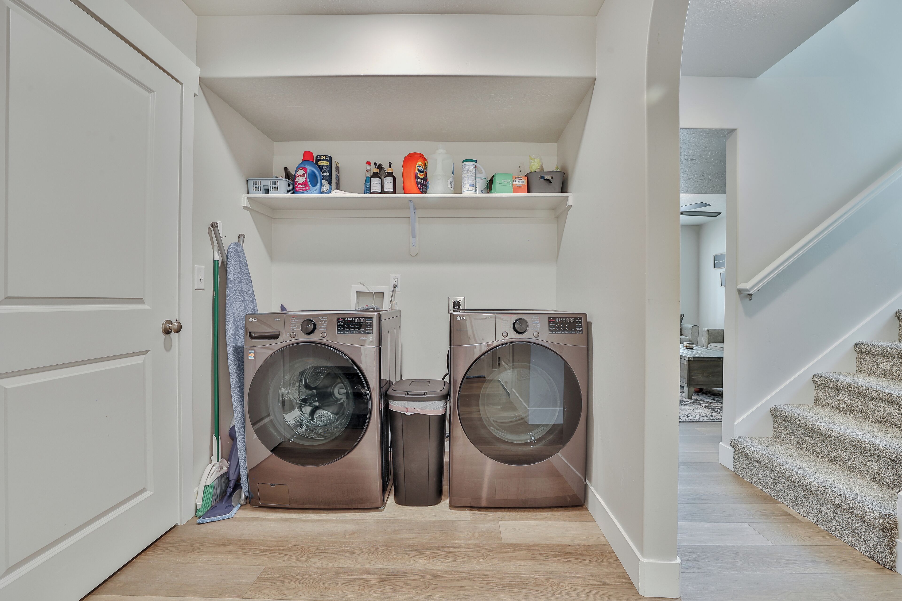 A laundry room with two front-loading washers and dryers, a shelf above with cleaning supplies, a broom, and a trash can.