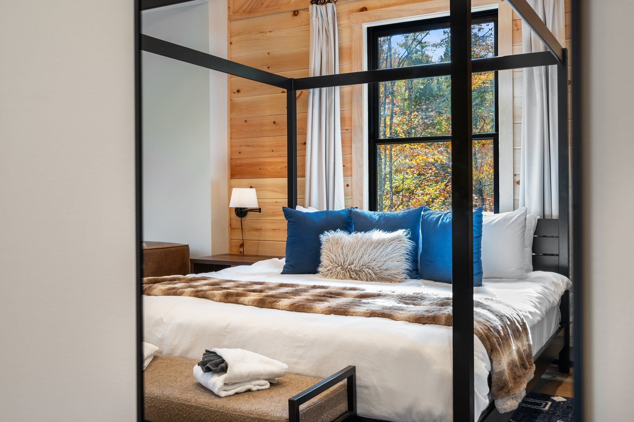 A four-poster bed with a fur throw, a cat on a cat tree, and a view of autumn trees through a large window.