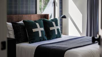 Een net opgemaakt bed met donkergroene kussens en een wit kruis motief, een houten hoofdbord en uitzicht op groen buiten door een raam.