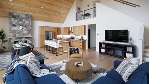 A spacious living area with a high ceiling, a stone fireplace, a kitchen with wooden cabinets, and a large TV on a black stand.
