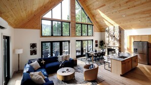 A spacious living area with a high ceiling, large windows, a stone fireplace, and a modern kitchen.