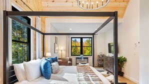 A modern bedroom with a large bed, a desk, and a view of trees through the window.