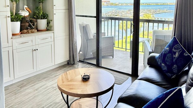 A modern living room with a sofa, a coffee table, and a view of a marina through large sliding glass doors. - Perrydise@ Pirates' Bay Top-Floor Studio with Panoramic Sound Views, Pools, Dock (Fort Walton Beach)