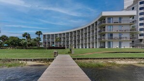 A modern hotel with a curved facade, multiple balconies, and a wooden dock leading to a water body. - Perrydise@ Pirates' Bay Top-Floor Studio with Panoramic Sound Views, Pools, Dock (Fort Walton Beach)