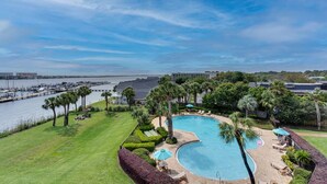A swimming pool with a curved edge, surrounded by palm trees and a grassy area. - Perrydise@ Pirates' Bay Top-Floor Studio with Panoramic Sound Views, Pools, Dock (Fort Walton Beach)