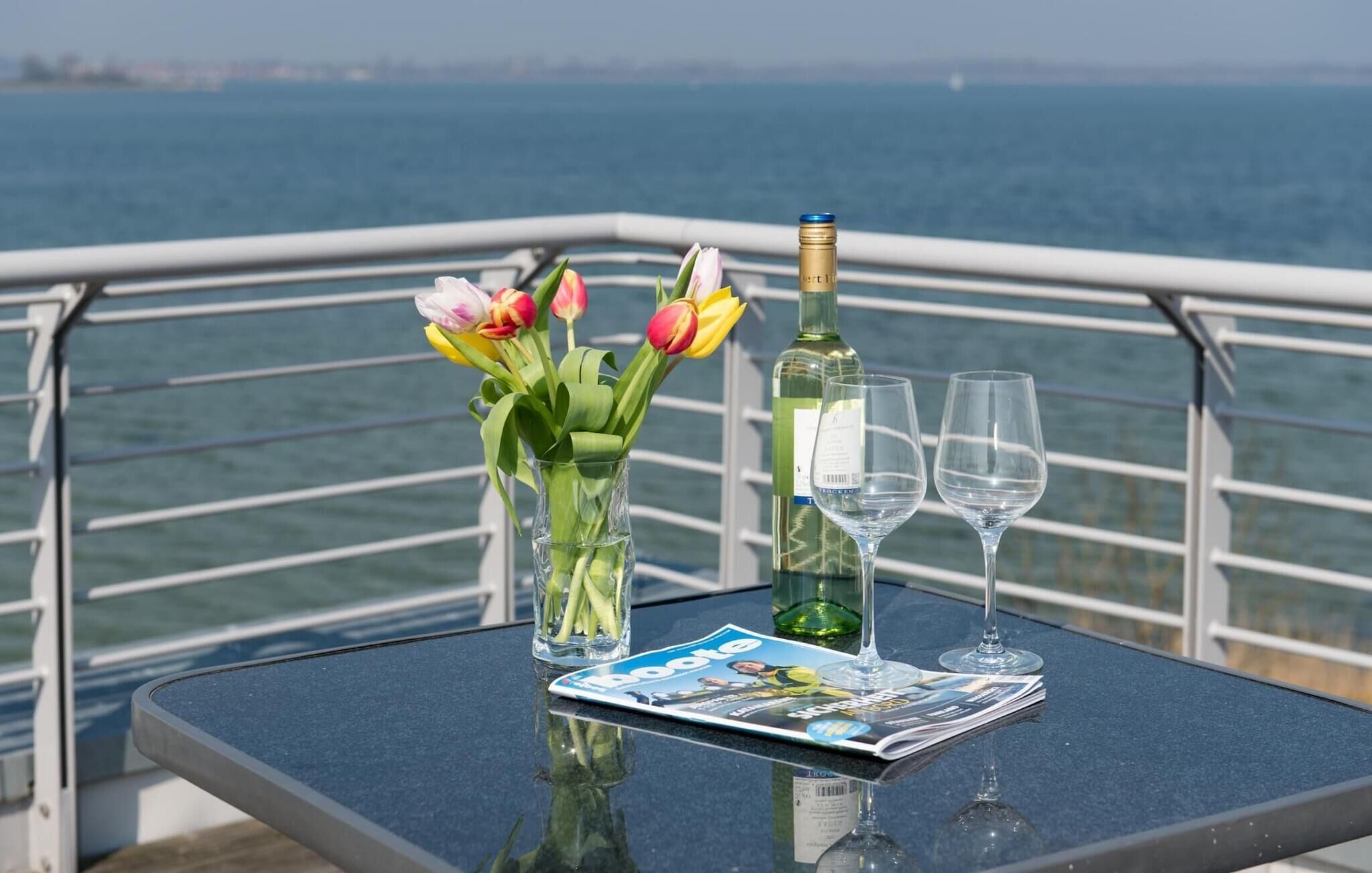 Ein Tisch mit einer Champagnerflasche, zwei Weingläsern, einer Zeitschrift und einem Strauß Tulpen auf einem Balkon mit Meerblick.