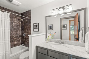Un bagno moderno con vasca, servizi igienici e lavabo. Alle pareti ci sono quadri incorniciati, uno specchio sopra il lavabo e una tenda da doccia.