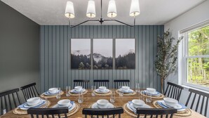 Comedor con mesa de madera dispuesta para seis personas, rodeada de sillas negras, una ventana grande en un lateral y un cuadro colgado en la pared.