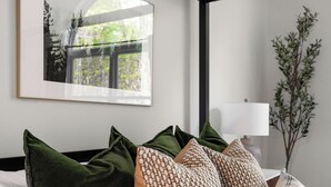 Un dormitorio ordenado con cama, almohadas, un cuadro enmarco, una lámpara y una planta.