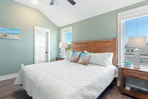 A bedroom with a large bed, wooden headboard, two bedside tables with lamps, and a view of the beach through the window.