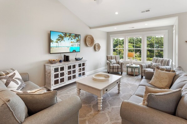 A spacious living room with a large TV, comfortable seating, and a view of the outdoors. - Coastal family escape w/ easy beach access & community pool (Kill Devil Hills)