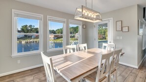 Zona pranzo con tavolo e sedie in legno, ampie finestre vista su un corpo d'acqua e vegetazione, e lampadario a sospensione.