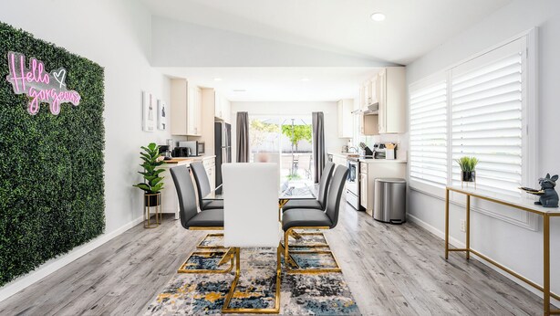 Una cocina moderna con armarios blancos, un comedor con una mesa y sillas de vidrio, y una pared verde con un letrero de neón.