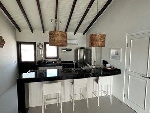 A modern kitchen with a black countertop, white bar stools, a stainless steel refrigerator, and a hanging light fixture. - Villa Veva fully equipped Waterfront Escape (Kralendijk)