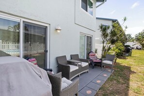 Un espace patio aménagé avec du mobilier d’extérieur, un barbecue couvert et une porte-fenêtre coulissante donnant accès à une piscine.