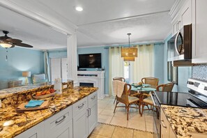 A modern kitchen with granite countertops, a dining area with wicker chairs, and a flat-screen TV above the fireplace.