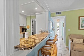 A kitchen with a marble countertop, wooden chairs, and a view into the dining area.
