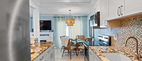 A modern kitchen with granite countertops, white cabinetry, a dining area with wicker chairs, and a tiled backsplash.