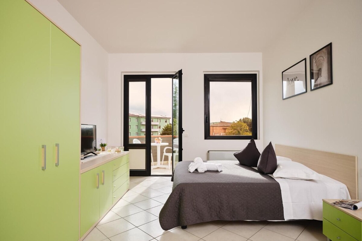 A hotel room with a bed, a green wardrobe, a dining area with a table and chairs, and a balcony with a view.