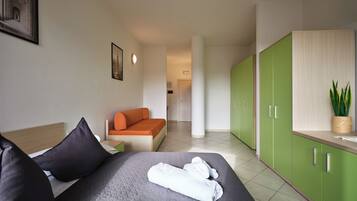 Une chambre d’hôtel avec un lit, une armoire verte, une petite table avec une plante, et un couloir menant à une autre chambre.