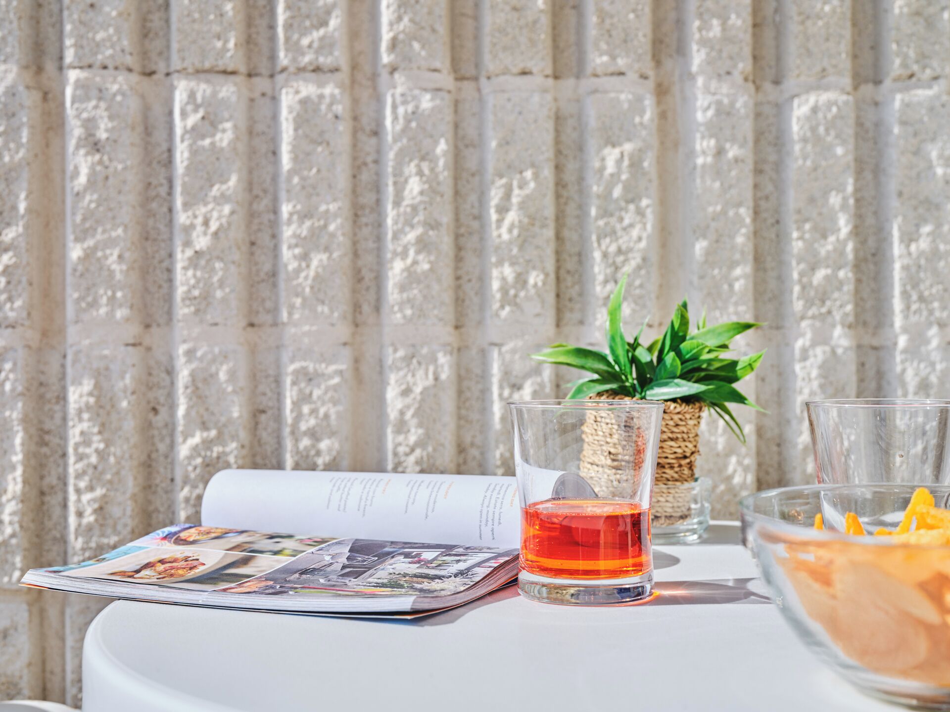 Une table avec un magazine, un verre contenant un liquide rouge, une petite plante en pot et un bol rempli de tranches d’orange.