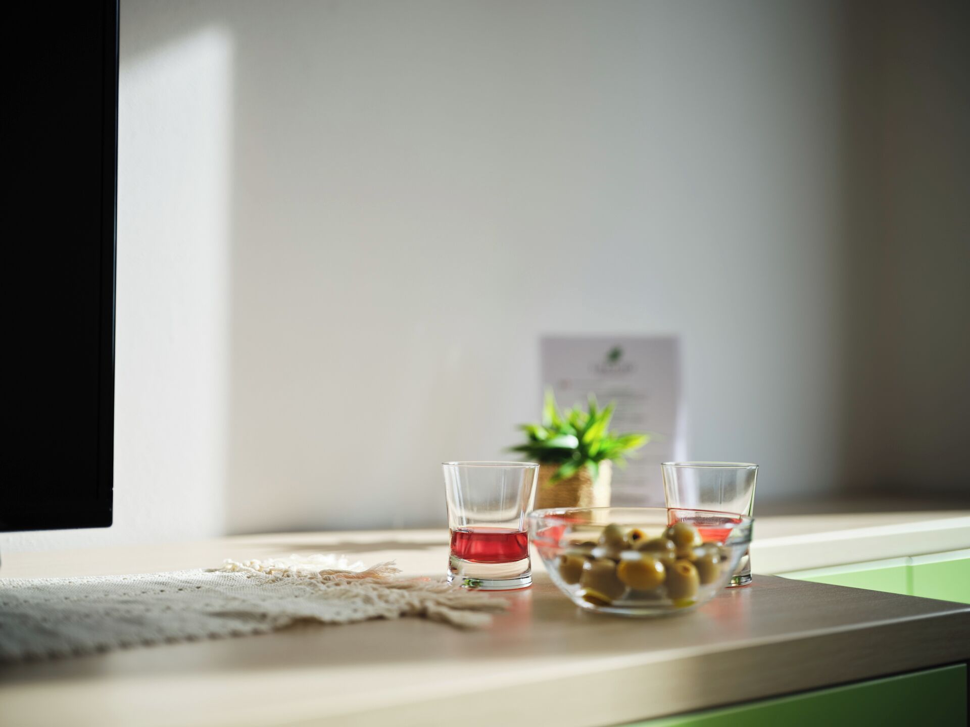 Una encimera de cocina con dos vasos de líquido rojo, un bol de aceitunas y una pequeña planta en maceta.