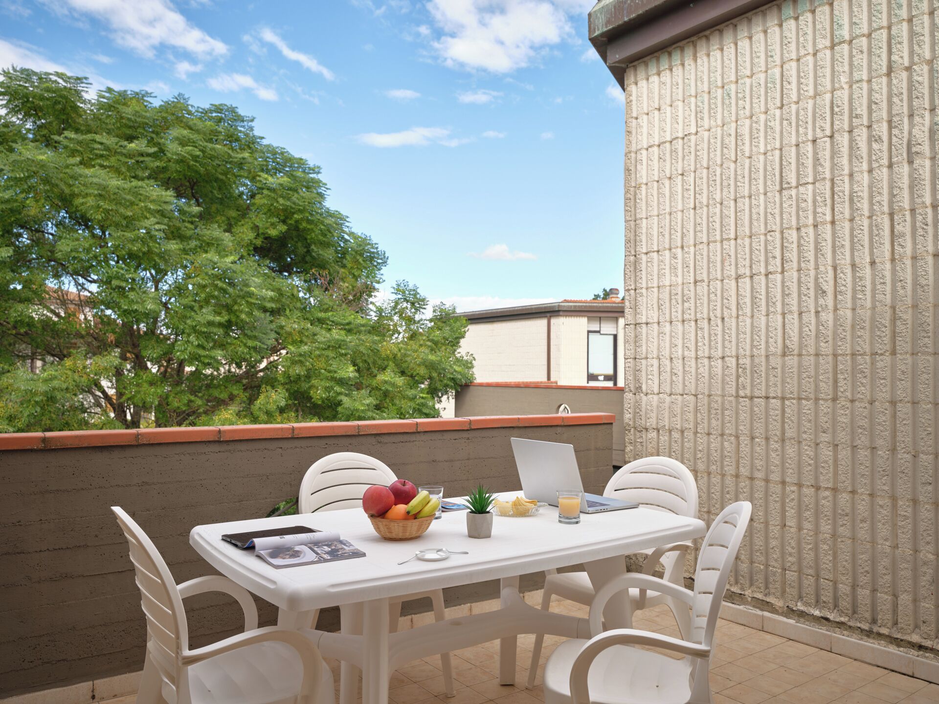 A white table with four chairs on an outdoor balcony, a basket of fruit, a laptop, and a glass of orange juice.