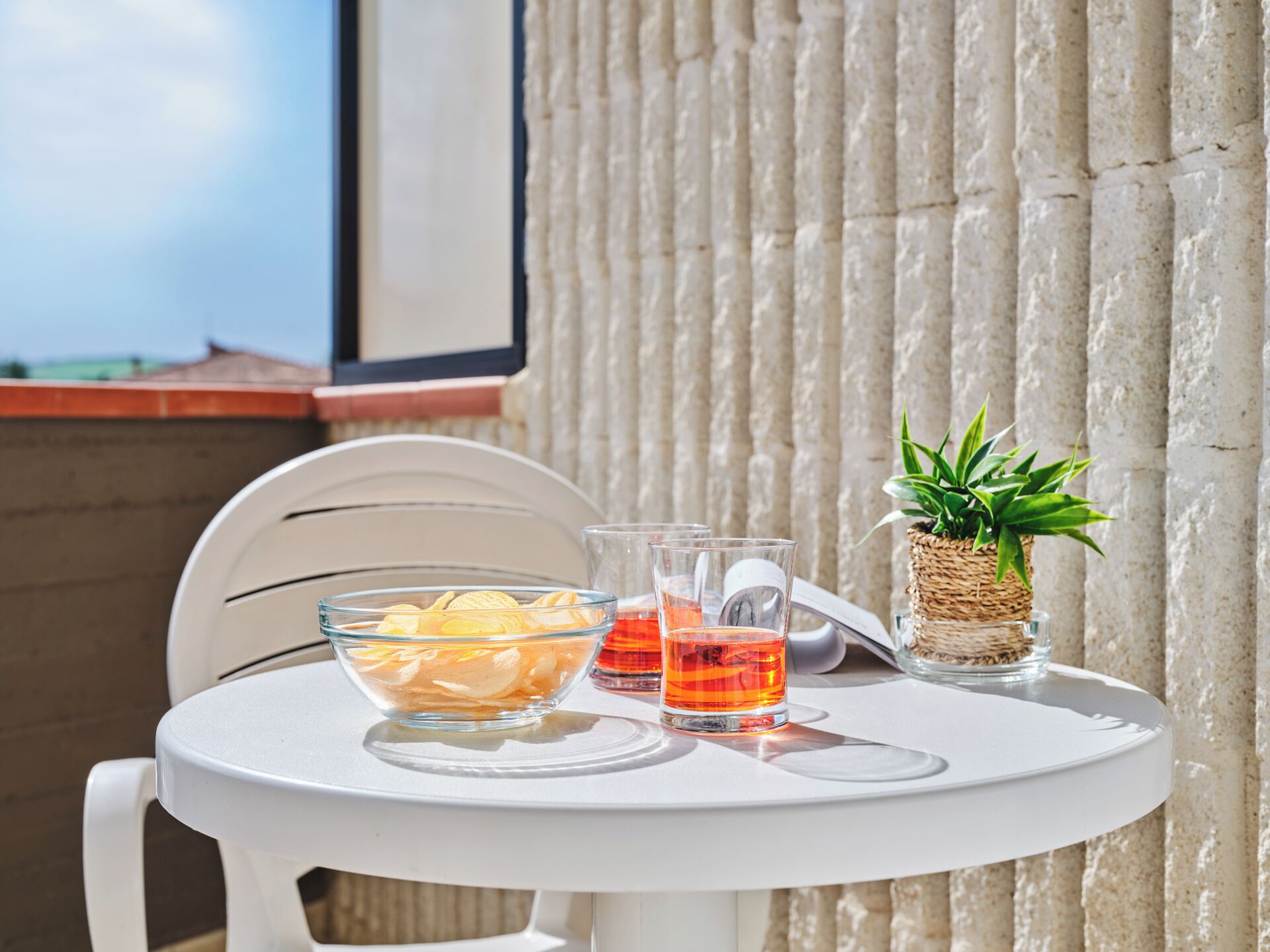 Una mesa blanca con un cuenco de aperitivos, dos vasos de jugo de naranja y una pequeña planta en maceta en un balcón.