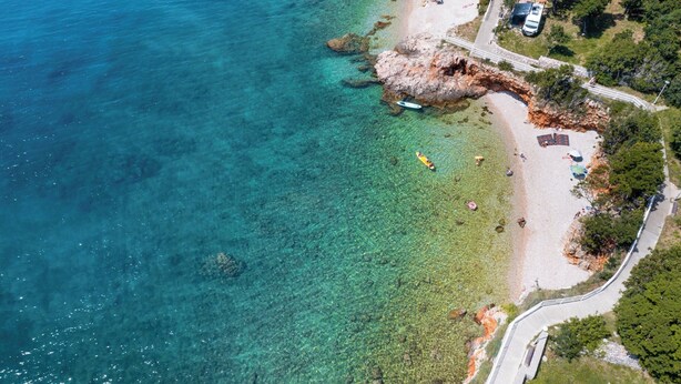 An aerial view of a coastal area with clear blue water, a sandy beach, and a rocky outcrop. There are people and umbrellas on the beach, and a road leading to the shore. - Villa Opera (Kvarner - Povile) (Povile)