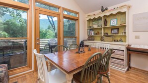 Un espace repas avec une table et des chaises en bois, une bibliothèque contenant divers objets et de grandes fenêtres donnant sur un paysage verdoyant.