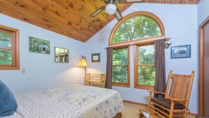 Une chambre à coucher avec un lit, un fauteuil à bascule, une fenêtre avec des rideaux et un ventilateur de plafond.