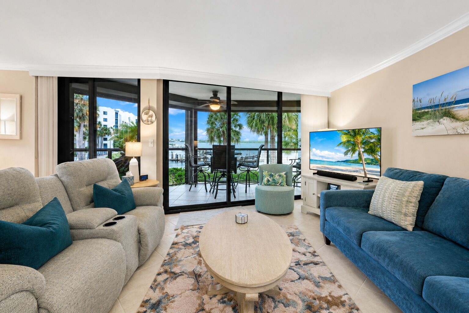A living room with a sofa, a coffee table, and a TV. There is a balcony with a view of the ocean and palm trees.