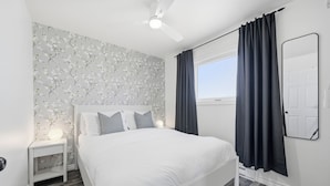 A bedroom with a white bed, a nightstand with a lamp, a ceiling fan, and a window with curtains. - The Birch (Prince Edward)