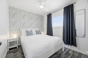 Un dormitorio con una cama blanca, una mesita de noche con lámpara, un ventilador de techo y una ventana con cortinas.