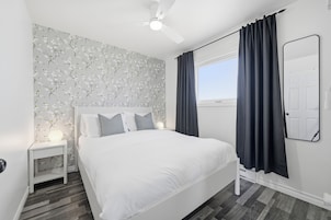 Un dormitorio con una cama blanca, una mesita de noche con lámpara, un ventilador de techo y una ventana con cortinas.