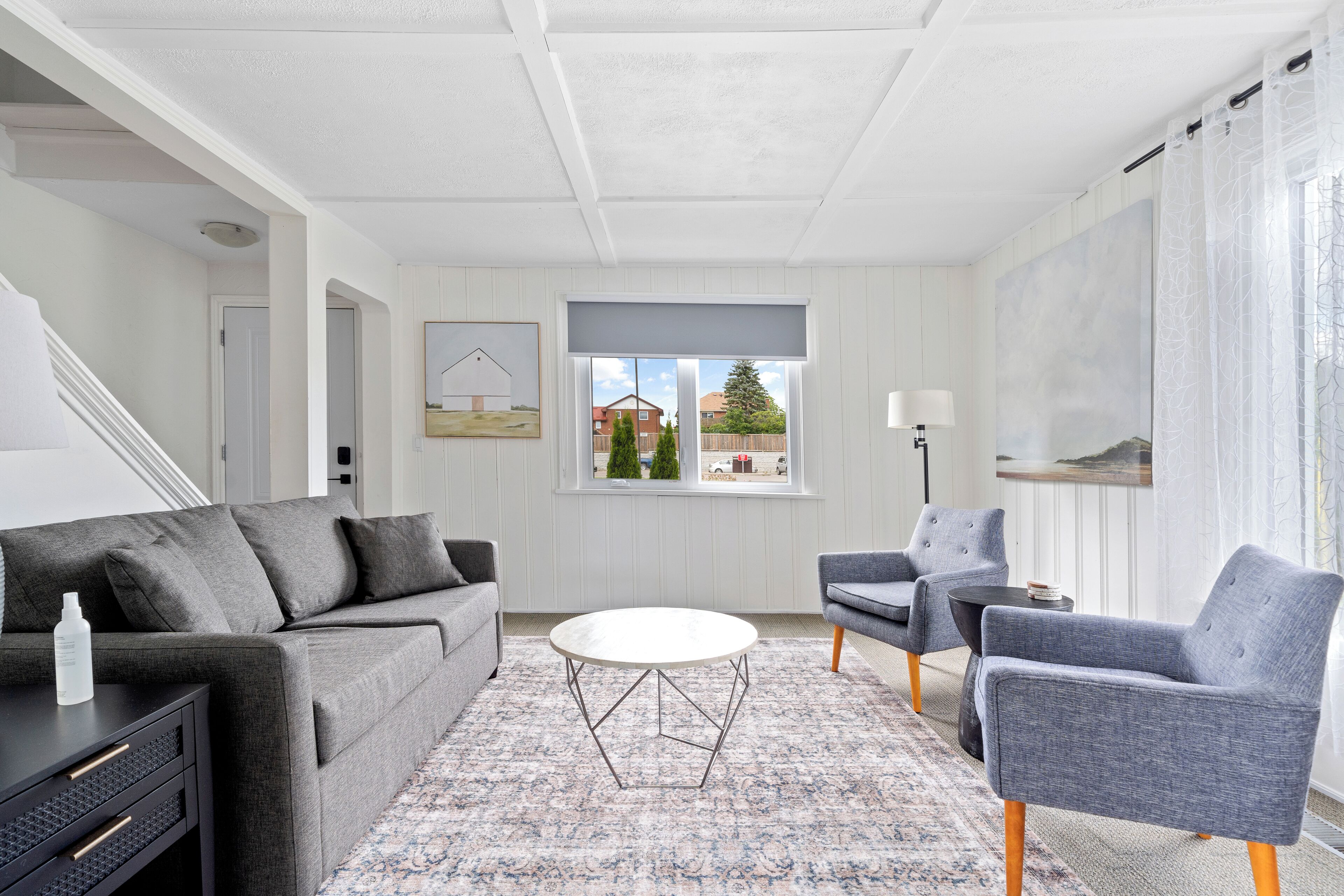 Una sala moderna con un sofá gris, una mesa redonda y dos sillones. Se ve una ventana con vista a un árbol y un edificio.