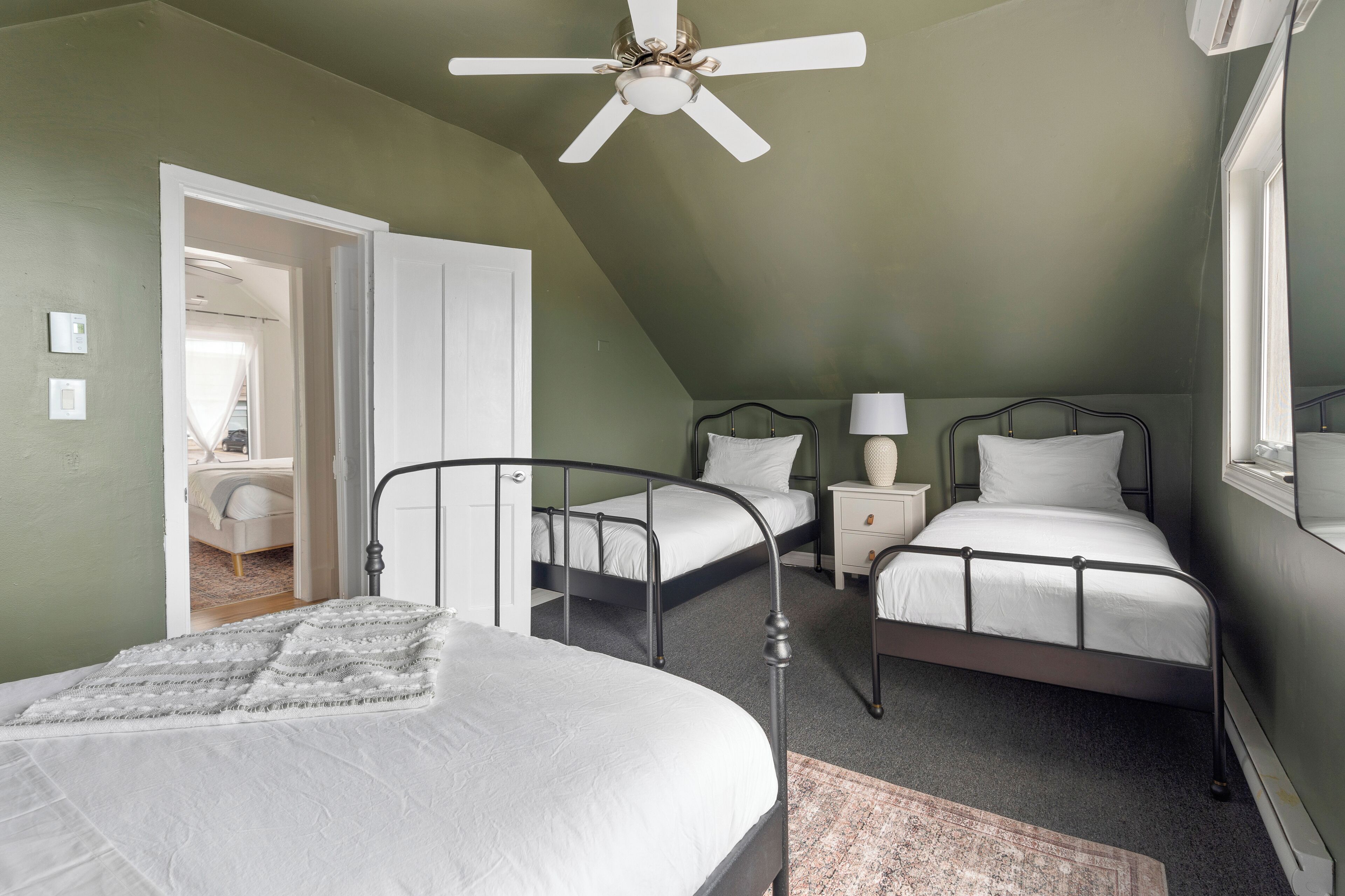 A bedroom with two beds, a ceiling fan, a window, and a door leading to another room.