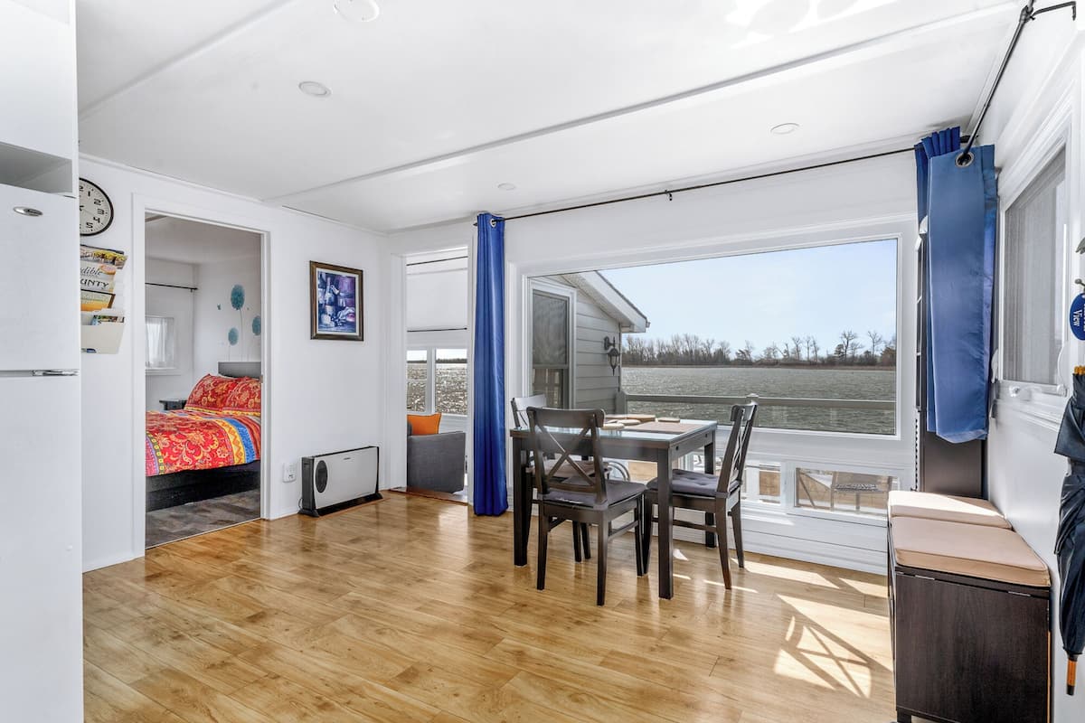 A room with a bed, a table with chairs, a window with a view of the water, and a clock on the wall.