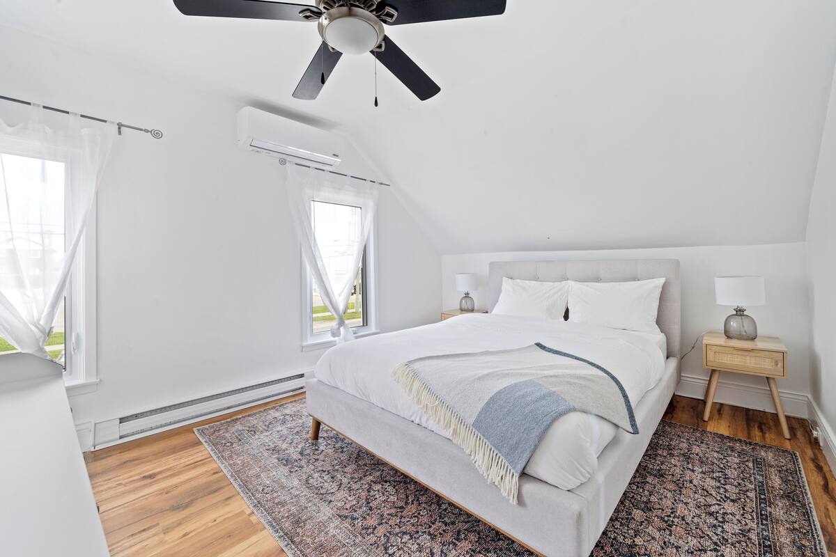 A bedroom with a bed, a ceiling fan, a nightstand, a lamp, and a window with curtains.