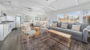 A spacious living area with a grey sofa, wooden coffee table, and light wood chairs. A kitchen with white appliances is visible in the background. - The Birch (Prince Edward)