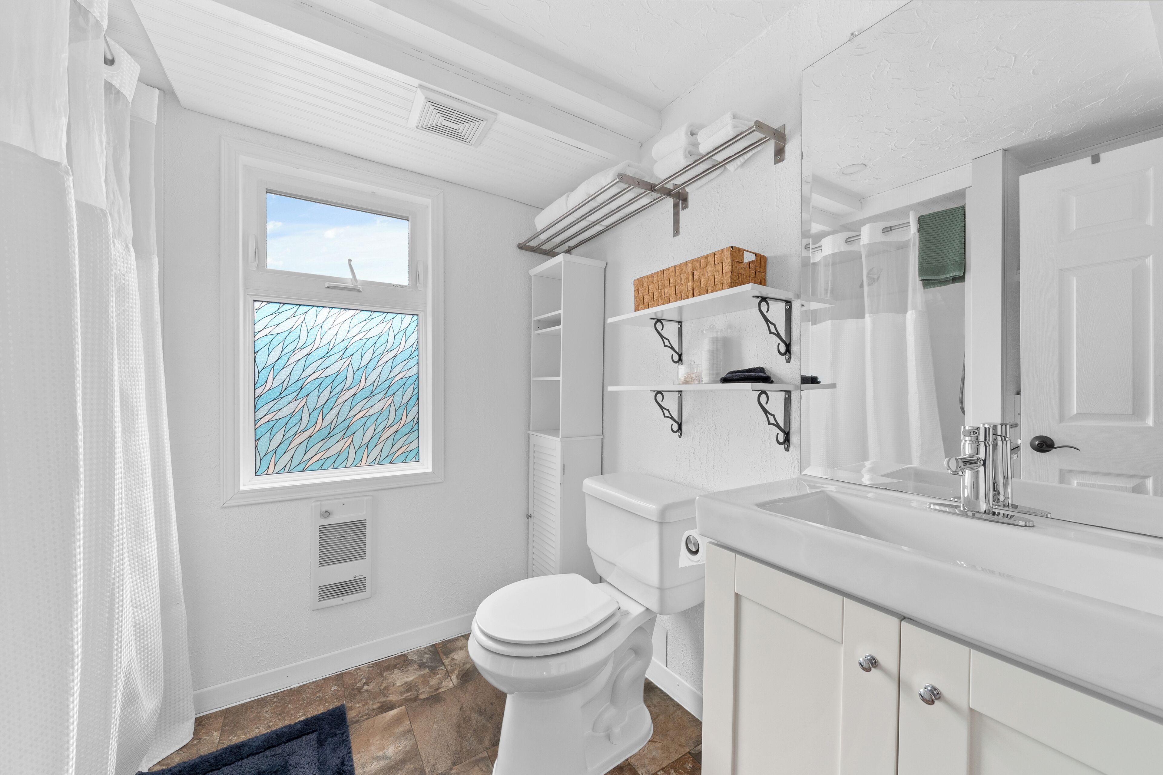 A modern bathroom with a toilet, sink, and shower. There is a window with a leaf-patterned view, shelves with toiletries, and a towel rack.