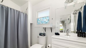 A bathroom with a white toilet, a shower curtain, a window with a floral design, a sink with a mirror, and toiletries on the counter. - The Birch (Prince Edward)