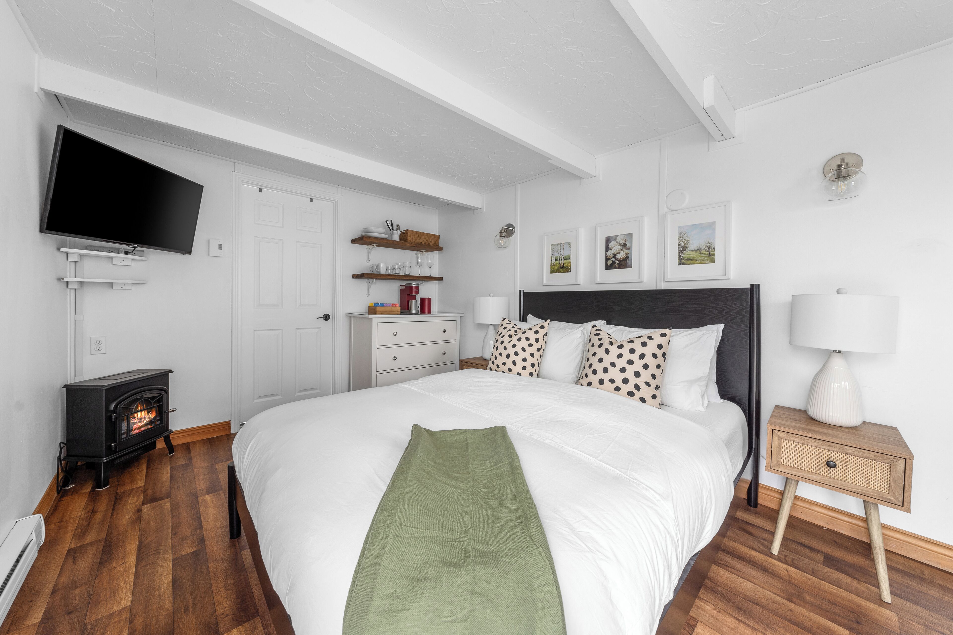 Un dormitorio con cama, televisor montado en la pared, pequeño calentador, mesita de noche con lámpara y otro mueble auxiliar.