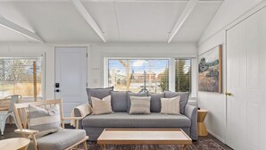 A modern living room with a grey sofa, wooden armchairs, a wooden coffee table, and a view of a residential area through large windows. - The Birch (Prince Edward)